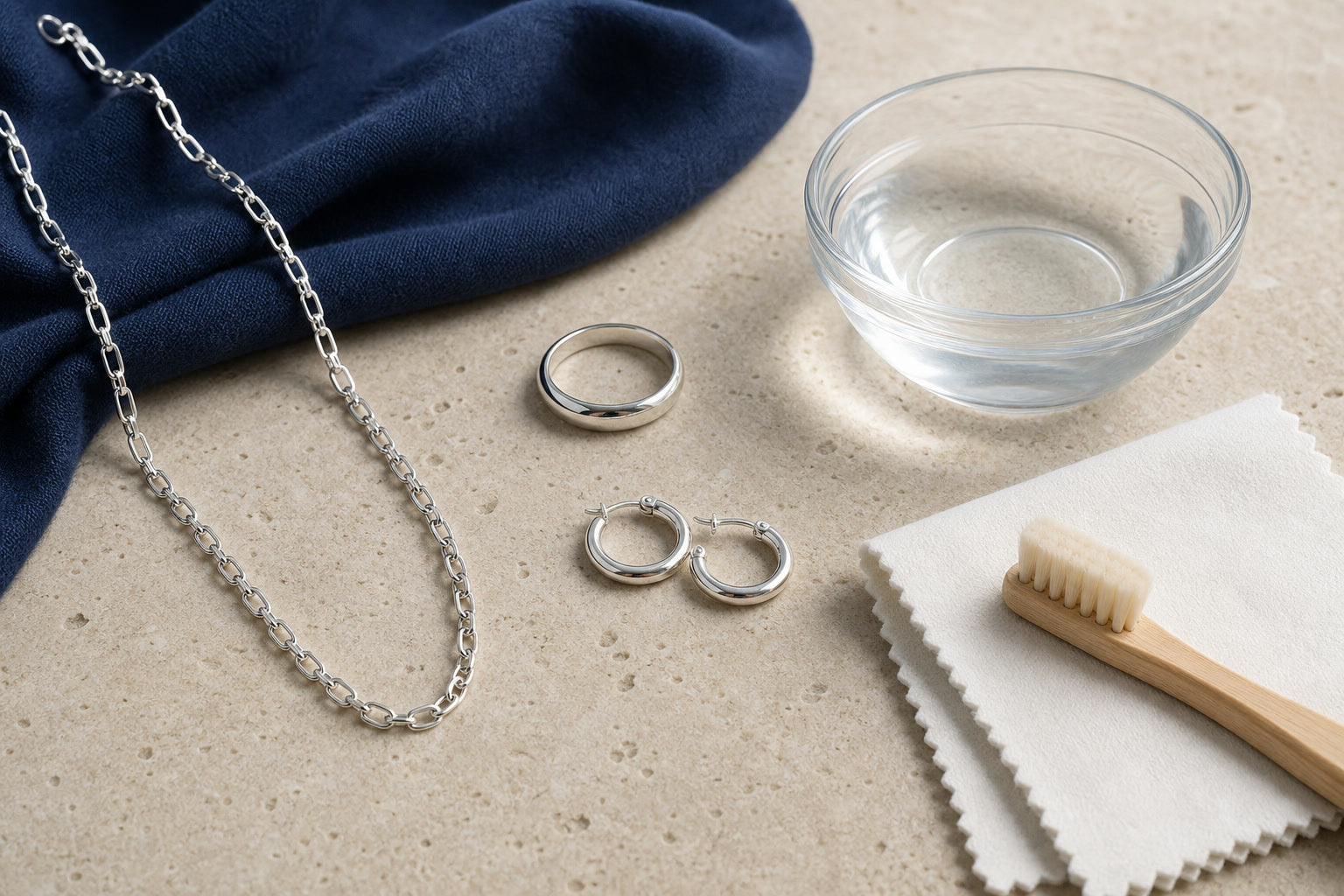 A sterling-silver chain, ring, and hoop earrings beside a soft cloth, clean water, and gentle brush